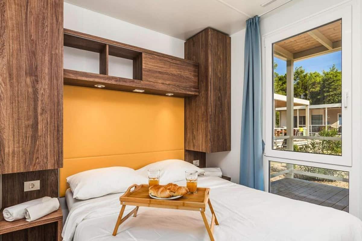 Une chambre d’hôtel avec un lit, une table de chevet en bois sur laquelle est posée une serviette, un plateau avec des croissants et du jus de fruits, et une fenêtre donnant sur l’extérieur.