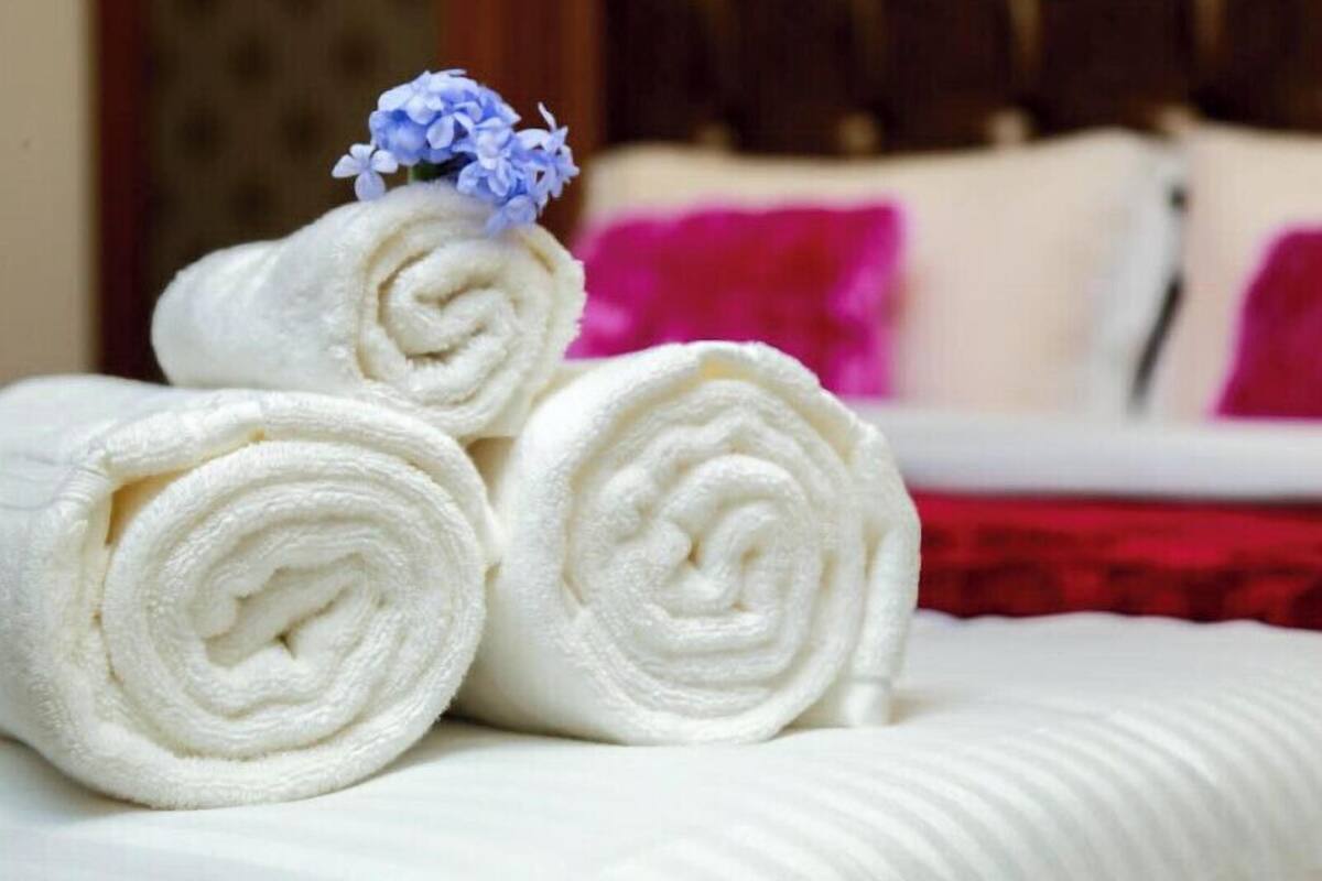 Three neatly rolled white towels placed on a bed with a white headboard and a red accent pillow.