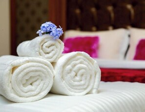 Three neatly rolled white towels placed on a bed with a white headboard and a red accent pillow. - Hotel S K Executive (Rajgurunagar)