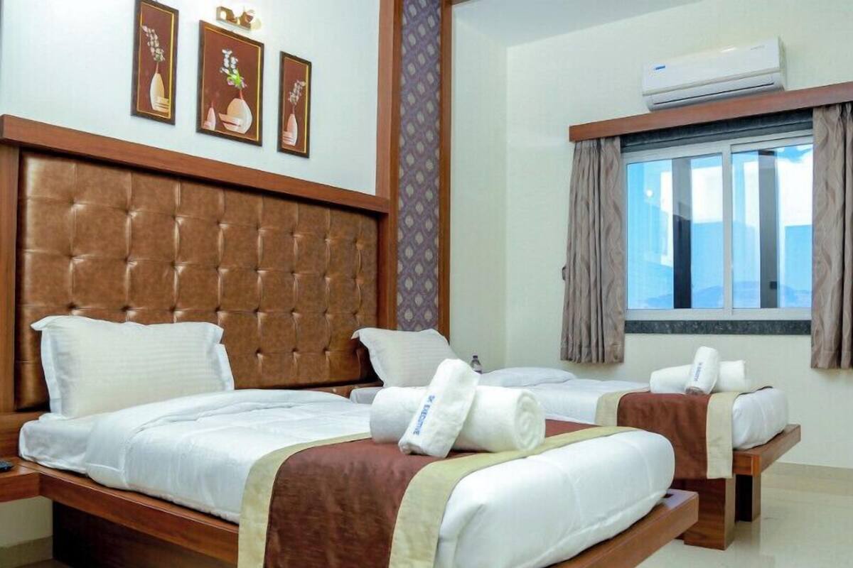 A hotel room with a large bed, two smaller beds, a window with curtains, and wall-mounted air conditioning.