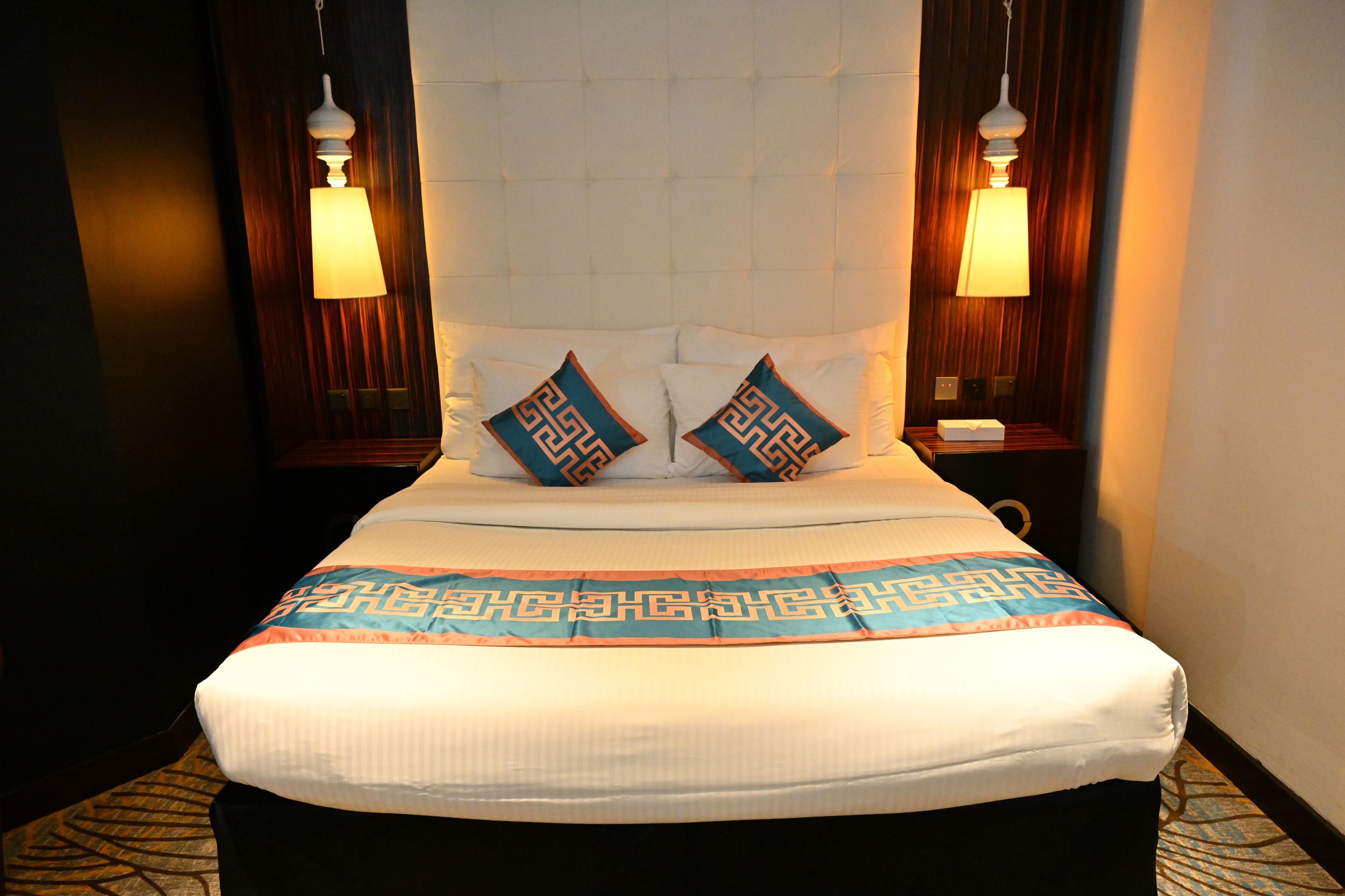 Habitación de hotel con una cama grande, dos almohadones decorativos, una colcha estampada y lámparas fijadas a la pared.