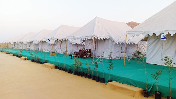 A row of white tents with numbered signs, lined up on a sandy surface with green tarps and potted plants in front. - Great Rann Resort (Bhuj)