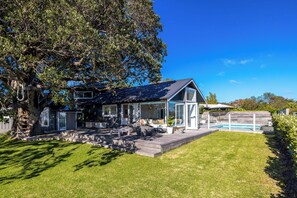 A house with a covered porch, a swimming pool, and a large tree in the yard. - Palm Beach Retreat - Now open for Christmas and New Year Booking! (Waiheke Island)