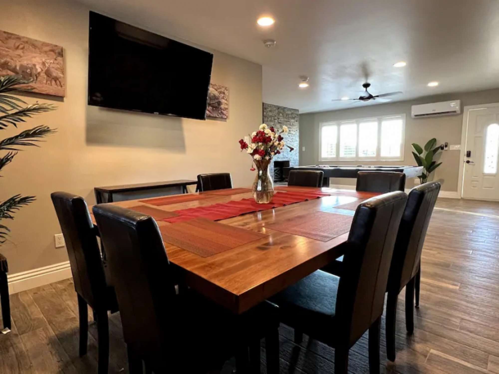 A dining area with a wooden table, black chairs, a vase with flowers, and a flat-screen TV mounted on the wall.