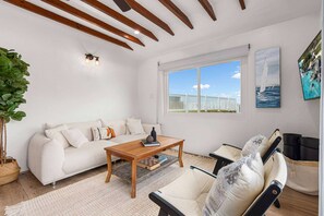 Un salon comprenant un canapé, une table basse et un fauteuil, une fenêtre donnant sur une clôture blanche, et un tableau représentant un voilier accroché au mur.