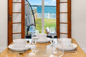 A dining table with white dishes and wine glasses, a wooden table, and a view of a body of water through open wooden doors. - New Luxury Oceanfront Unit with Private Beach (Southampton)