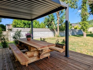 Une terrasse en bois avec une table de pique-nique et des bancs, entourée d’un jardin et d’une clôture blanche.