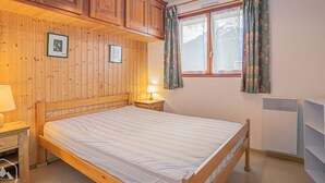 Een kamer met houten lambrisering, een bed, nachtkastje, lamp, raam met gordijnen en een radiator.