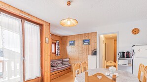 Een gezellige kamer met een houten bankje, een eettafel met stoelen, een koelkast en een klok aan de muur.