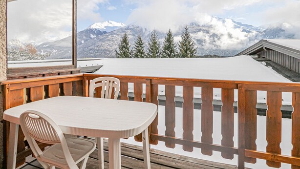 Een balkon met een houten hekwerk, een witte tafel en twee stoelen. Een groot raam biedt uitzicht op besneeuwde bergen en bomen.