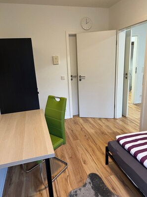 A room with a wooden floor, a bed with a striped bedspread, a green chair, a desk, and a white door with a clock. - Apartment 'An Littard Kull' with Private Terrace and Wi-Fi (Neukirchen-Vluyn)