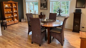 Un comedor con mesa de madera, sillas de mimbre, un armario con vajilla y dos ventanas con persianas.