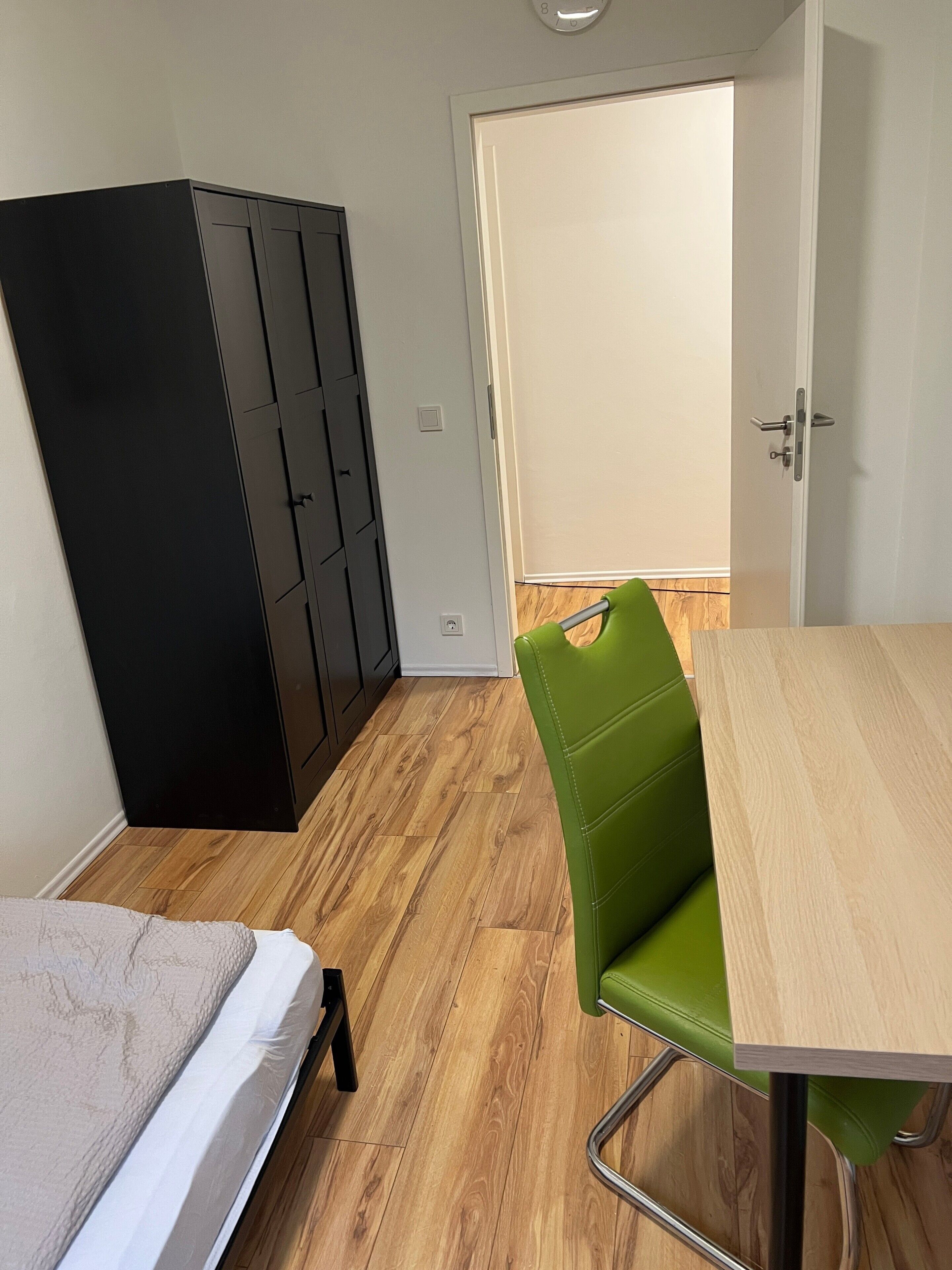 Huoneessa on sänky, vihreä tuoli, puinen pöytä, musta vaatekaappi ja ovi.