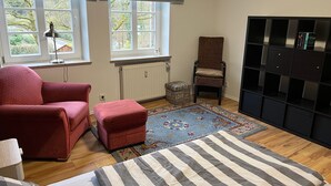 Una habitación con una cama, un sillón rojo, un pequeño taburete, una silla, una estantería y una ventana con persianas.