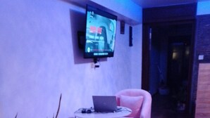 A room with a flat-screen TV mounted on the wall, a laptop on a small table, two pink chairs, and a potted plant.