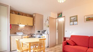 Een functionele keuken met houten kastjes, een eettafel met stoelen, een rode bank en een klein zithoekje met een televisie.