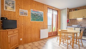 Een kamer met houten lambrisering, een eettafel met stoelen, een keukenhoek en een televisie op een kast.