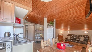 Cocina compacta con lavamanos, estufa y refrigerador. Hay una mesa de comedor con sillas y un tazón rojo. La habitación tiene paneles de techo de madera y una cama litera al fondo.