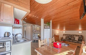 Une cuisine compacte Ă©quipĂ©e dâun Ă©vier, dâune cuisiniĂšre et dotĂ©e dâun plafond en bois. On y trouve un coin repas avec une table et des chaises, ainsi quâun canapĂ© Ă lâarriĂšre-plan.
