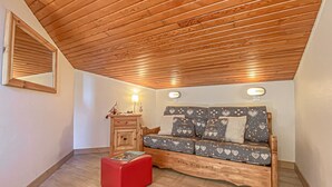 Una habitación acogedora con techo de madera, un sofá con cojines estampados con motivos de corazón, un pequeño mueble de madera y un reposapiés rojo.
