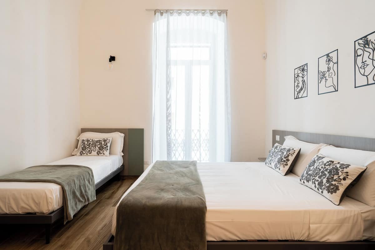 A room with two single beds, a bench, and a large window with sheer curtains.