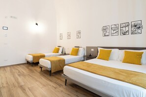 A hotel room with two single beds and one double bed, each with white bedding and yellow pillows. The room has a wooden floor and white walls. There are wall-mounted light switches and a small table with a chair. - Nicolaus 204 - Brown & Grey Rooms by Wonderful Italy (Bari)