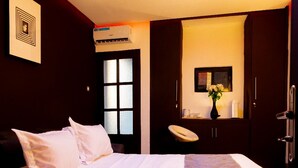 A hotel room with a bed, a vanity with a mirror, a vase with flowers, and a window with blinds. - The Pandora: Secure and stylish studio with sofa bed, WiFi, smart TV and tub (Yaoundé)