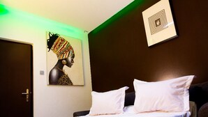 A hotel room with a bed, white pillows, a door, and a wall with a framed artwork. - The Pandora: Secure and stylish studio with sofa bed, WiFi, smart TV and tub (Yaoundé)