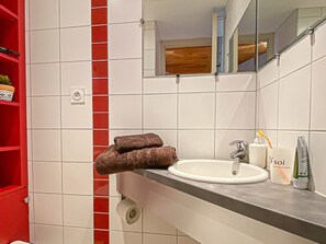 Ein Badezimmer mit einem weißen Waschbecken, einem Spiegel, einem Handtuchhalter mit zwei Handtüchern, Toilettenartikeln und einem roten Regal mit einer Topfpflanze.