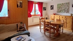 A cozy room with a wooden floor, a sofa, a dining table with chairs, a kitchen area, and a window with red curtains. - Cozy apartment near leisure, 6 people, Wifi (Aussois)
