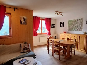 Ein gemütliches Zimmer mit Holzboden, einer Couch, einem Esstisch mit Stühlen, einer Küchenzeile und einem Fenster mit roten Vorhängen.