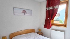 A bedroom with a wooden headboard, a single bed, a nightstand with a lamp, a window with a red curtain, and a wall-mounted picture. - Cozy apartment near leisure, 6 people, Wifi (Aussois)