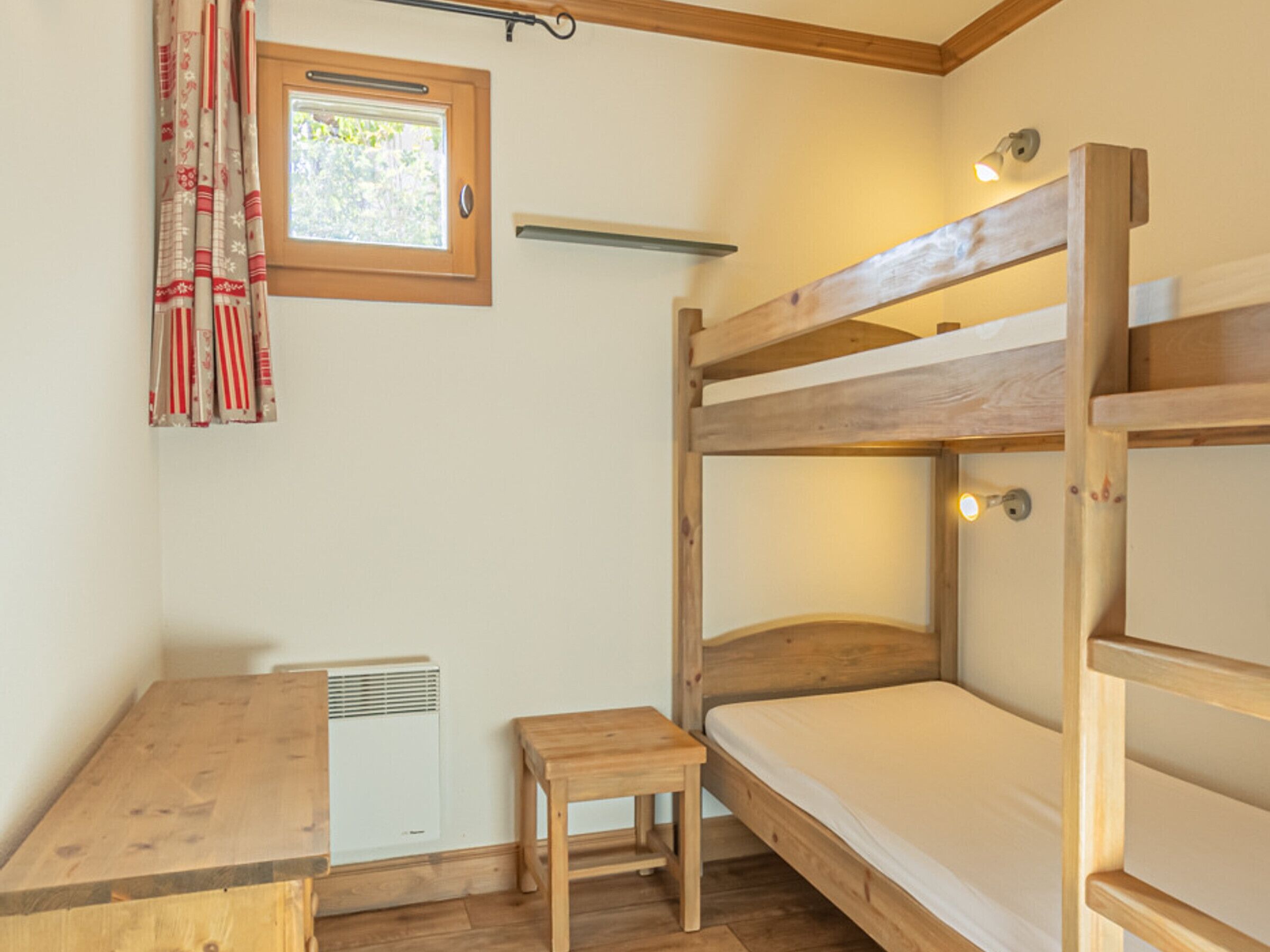 Een slaapkamer met een stapelbed, een houten bureau en stoel, een klein raam met gordijnen en een wandplank.