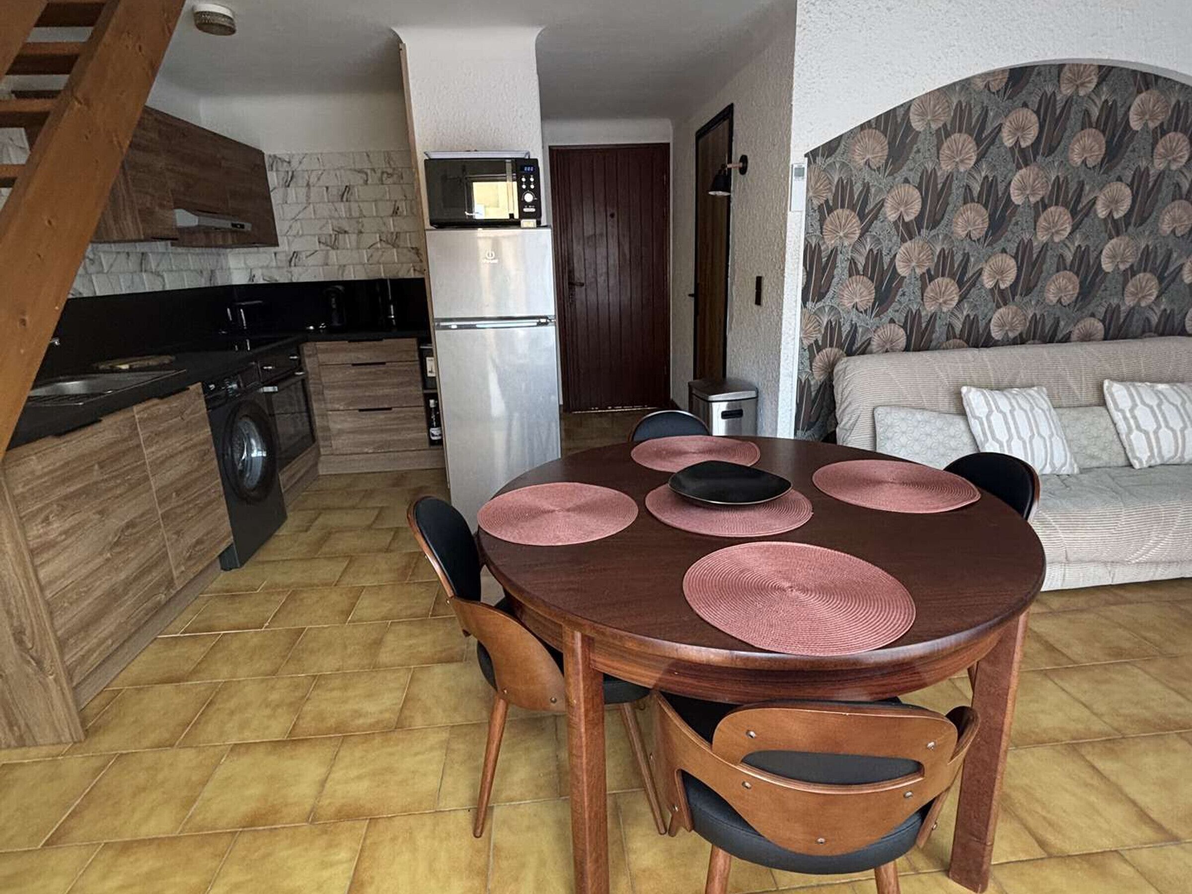 Ein Essbereich mit Holztisch und Stühlen, eine Küche mit Kühlschrank und Mikrowelle sowie ein Wohnzimmer mit Sofa und Wandpapier mit Muster.