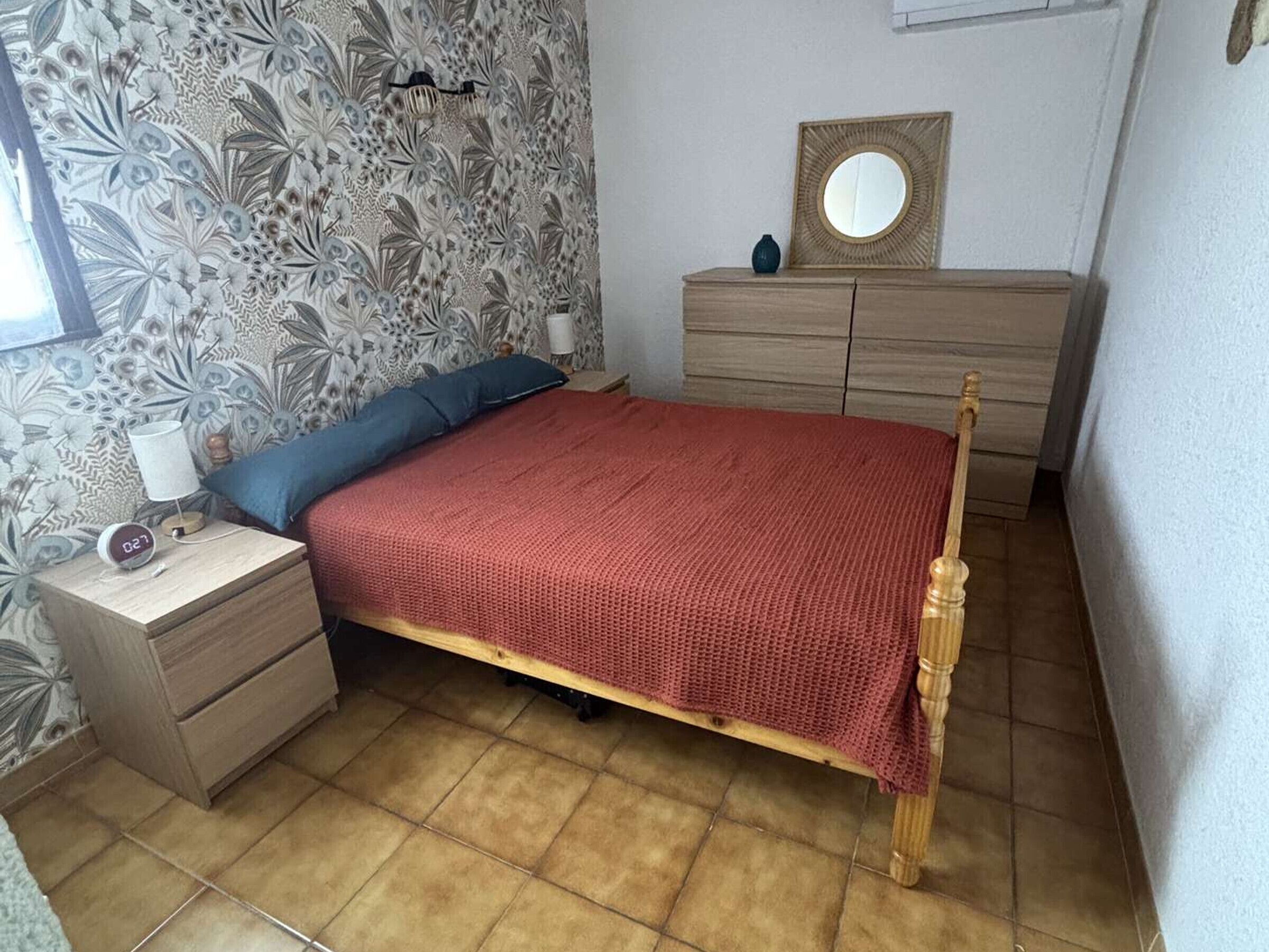 A bedroom with a wooden bed, a nightstand with a lamp, a dresser, and a mirror on the wall.
