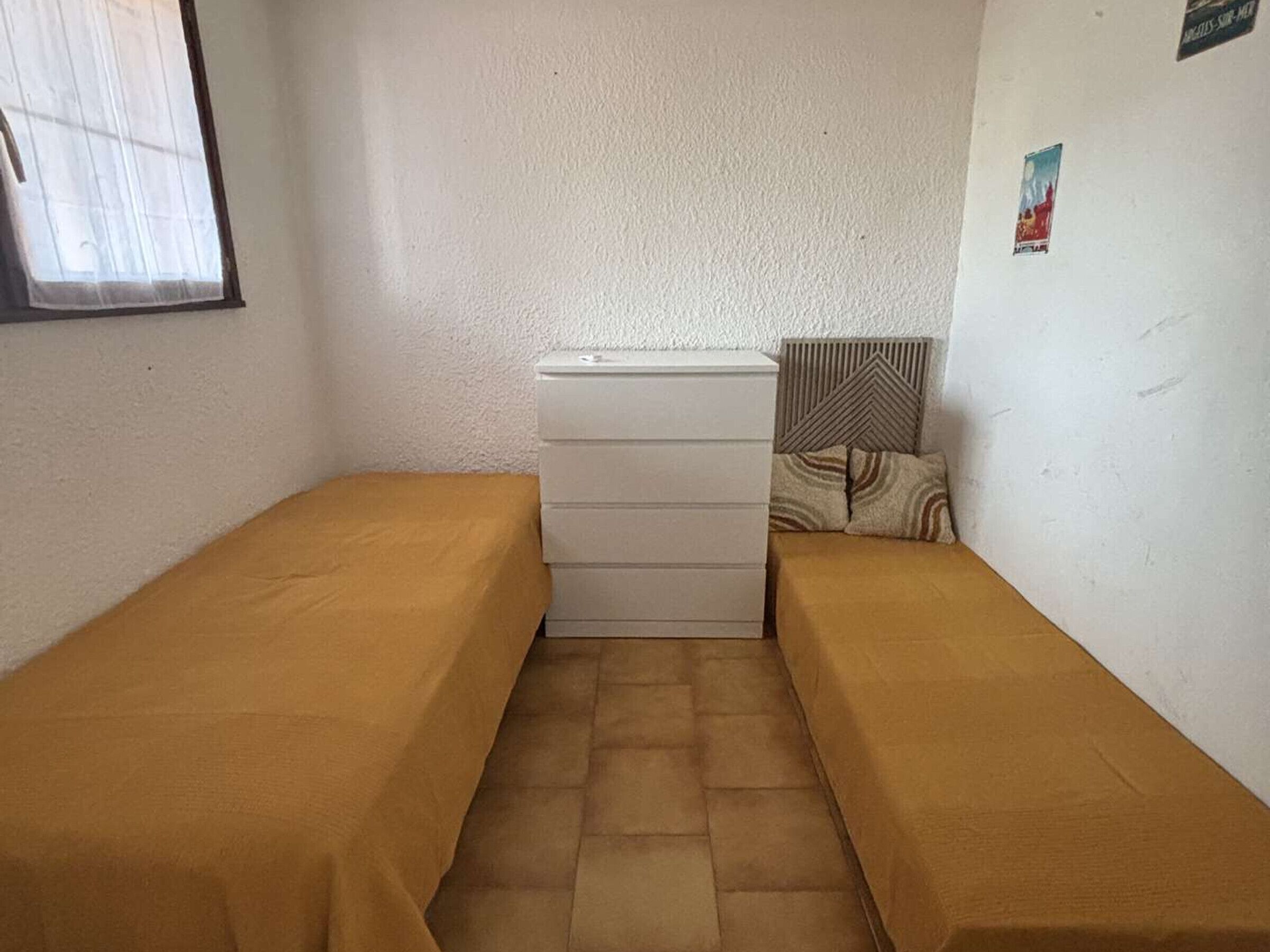 Een kleine kamer met twee eenpersoonsbedden, een dressoir, een radiator en een raam met dunne gordijnen.