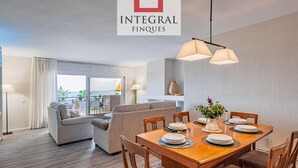 A modern dining area with a wooden table set for four, a sofa, and a view of the sea through large windows. - Beautiful sea-view apartment with WiFi and parking in Palamós (Palamós)
