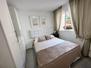 Quarto com cama, mesa de cabeceira, janela com cortinas e um quadro na parede.
