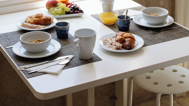 Una mesa de desayuno con un tazón de cereal, un croissant, un plato de fruta, una taza y un vaso de jugo de naranja.