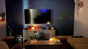 A living room with a large flat-screen TV mounted on a dark wall with star patterns, a wooden coffee table with candles, a small plant, and a pumpkin decoration. - Charming, Private , 3BR, 1Bath Main Floor Close to all action Stadiums & Sports (Minneapolis)