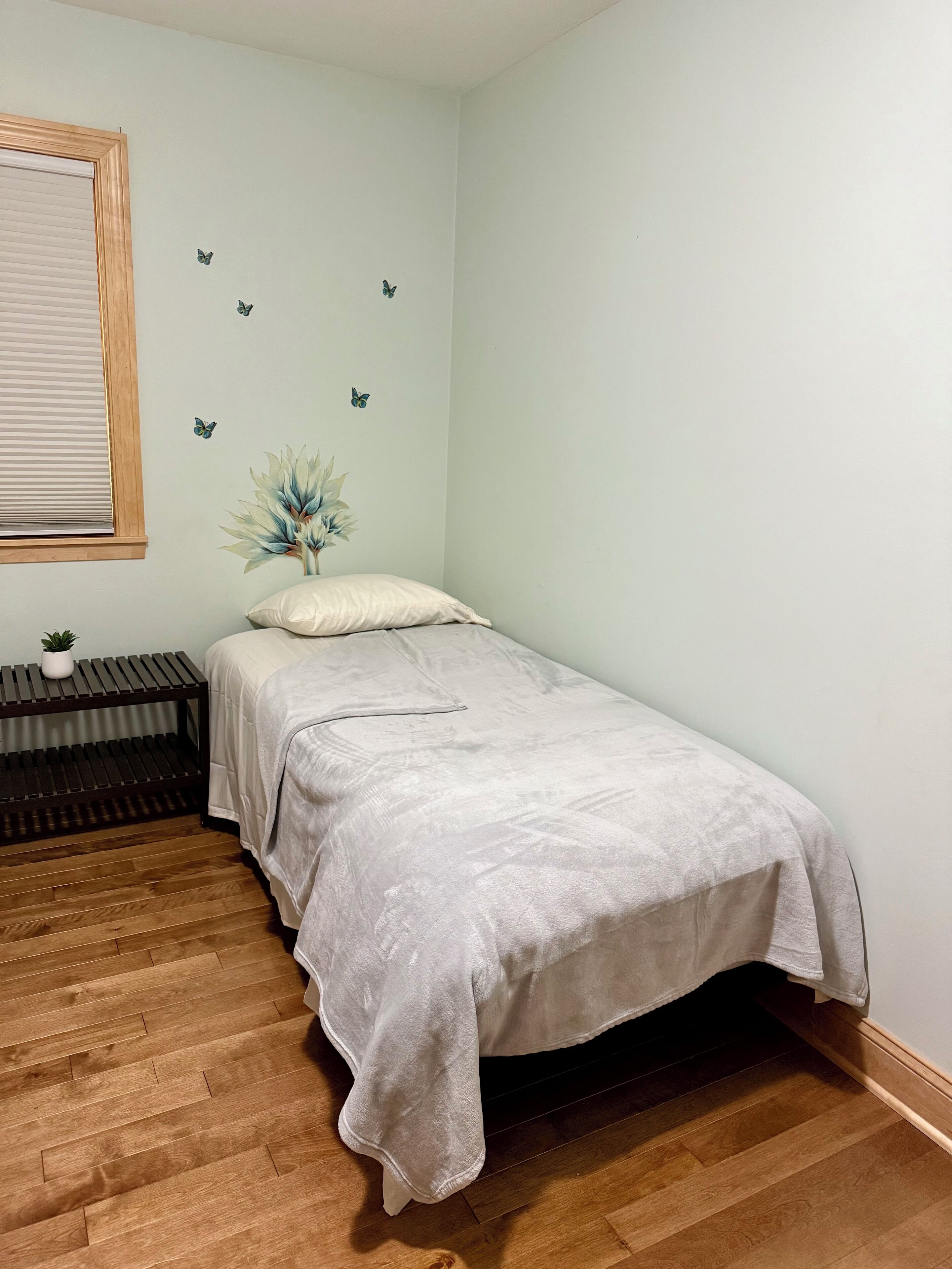 Un dormitorio con cama, mesita de noche, ventana con persianas, suelo de madera y decoración en las paredes, incluyendo mariposas y una planta.