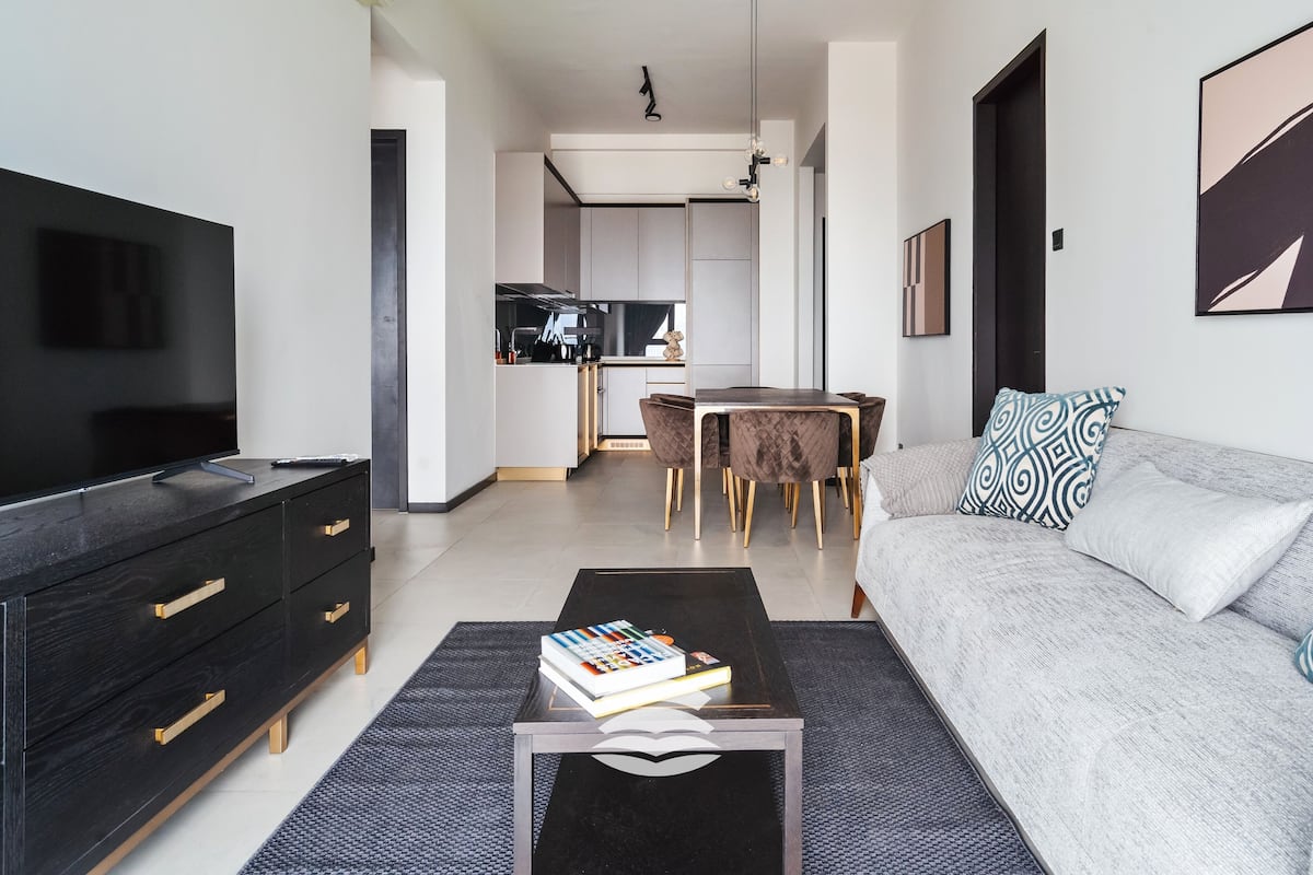 A modern living room with a flat-screen TV, a sofa, a coffee table with books, and a dining area with a table and chairs.