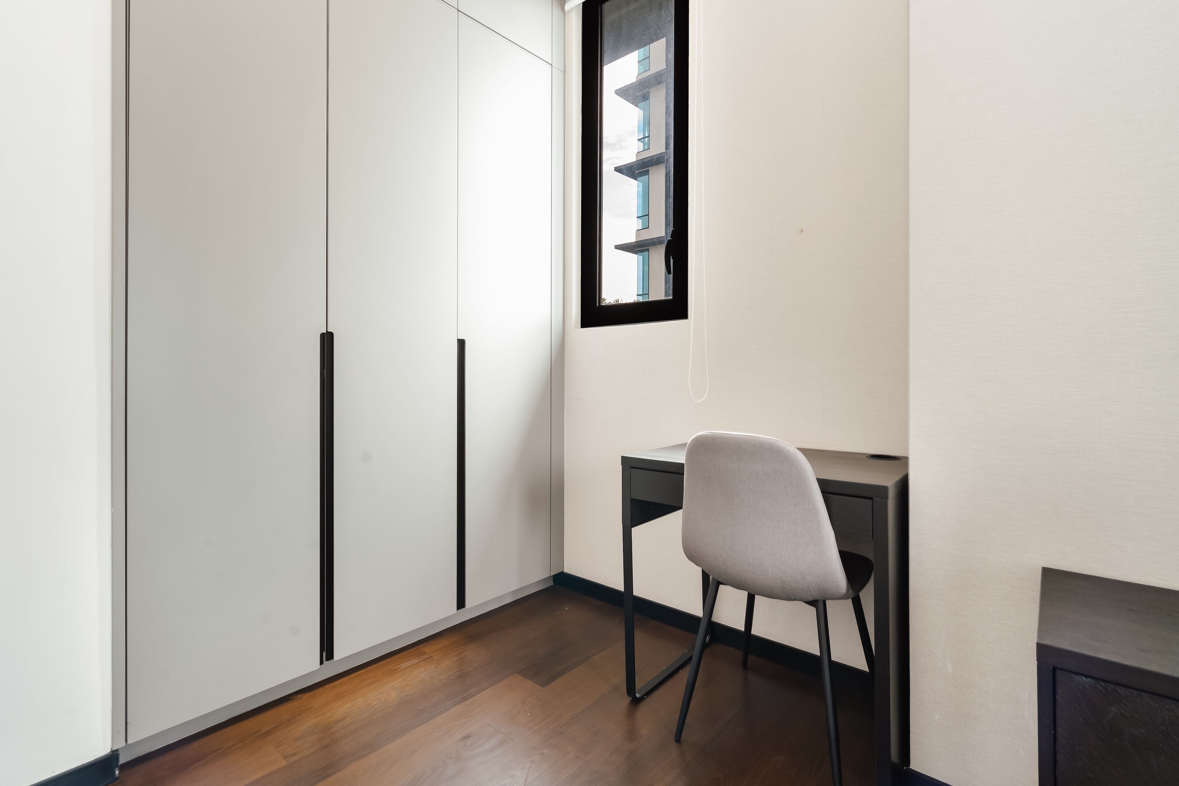 A room with a desk and chair, a wardrobe, and a window with a view of a building.