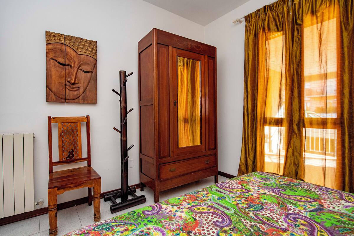 Ein Schlafzimmer mit einem Holzschrank, einem Sessel, einer farbenfrohen Bettdecke und einem Buddha-Gemälde an der Wand.
