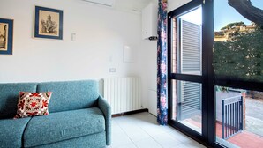 A living room with a teal sofa, a radiator, and a large window with a view of a building and trees. - LA TORRE Appartamenti (Castiglione della Pescaia)