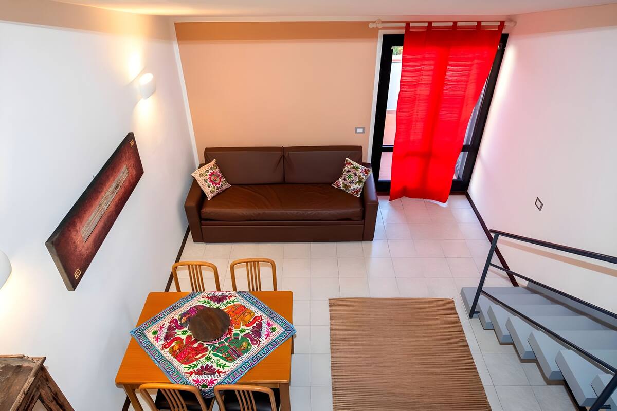 Ein Wohnzimmer mit einer Couch, einem Esstisch mit Stühlen, einer Treppe und einem roten Vorhang.