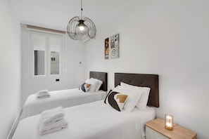 Quarto de hotel com duas camas, uma mesinha de cabeceira com abajur, uma janela com persianas e uma obra de arte pendurada na parede.