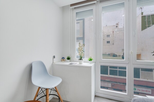 A corner room with a white radiator, a blue chair, a small table, a window with a view of buildings, and a potted plant.