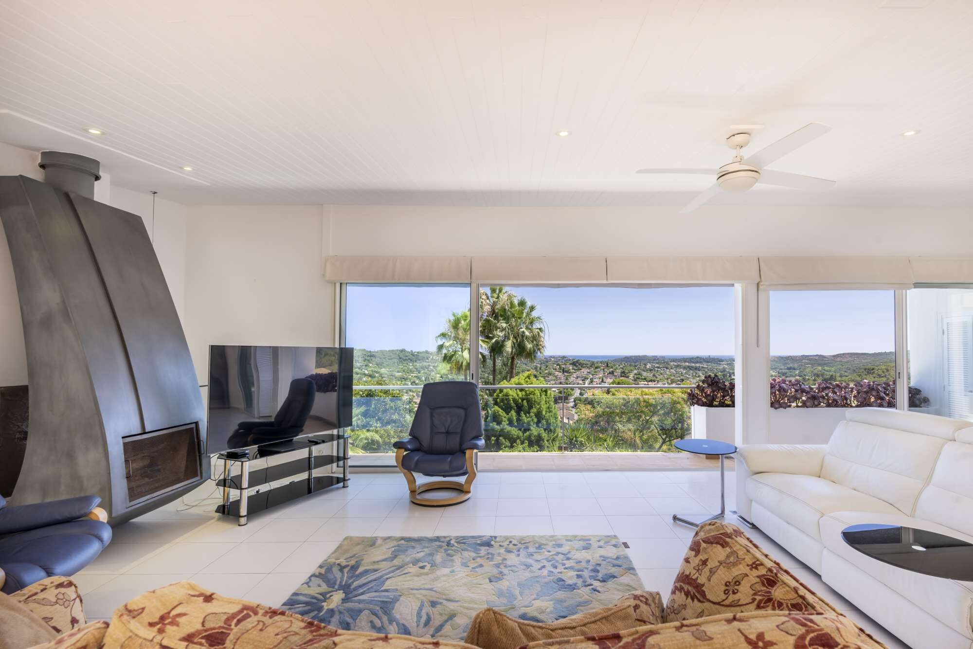 Ein modernes Wohnzimmer mit Fernseher, Sofa und Sesseln, große Fenster mit Blick auf Grünflächen und Gebäude.