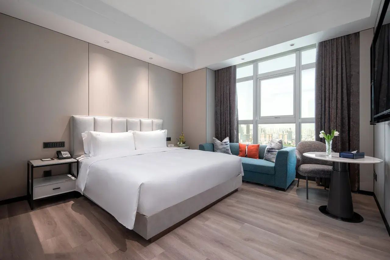 Habitación de hotel moderna con una cama grande, zona de estar con sofá y sillones, una mesita redonda y un ventanal con vistas a la ciudad.
